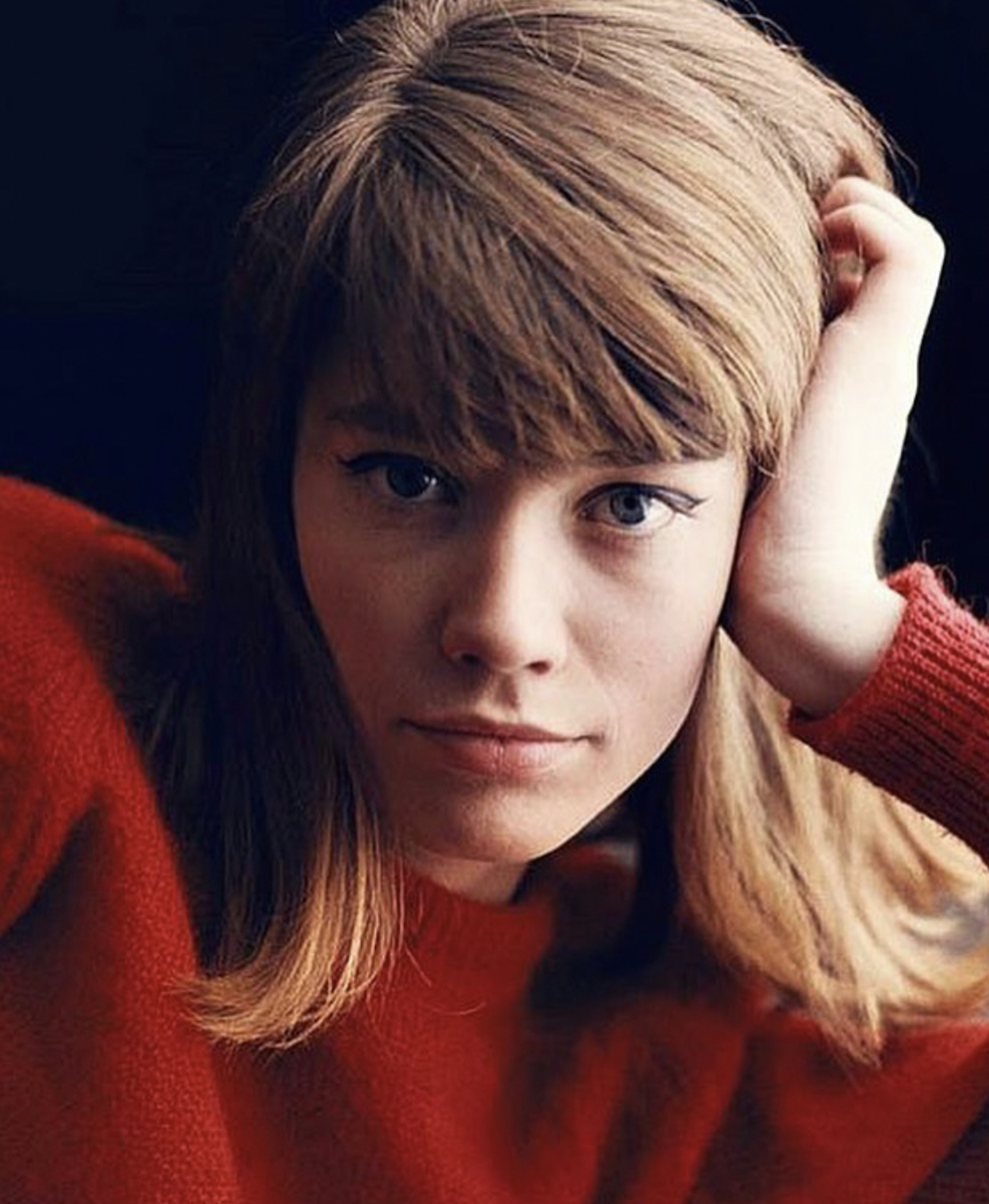 Close-up portrait of a young woman with light brown hair and a red sweater, resting her head on her hand against a dark background.