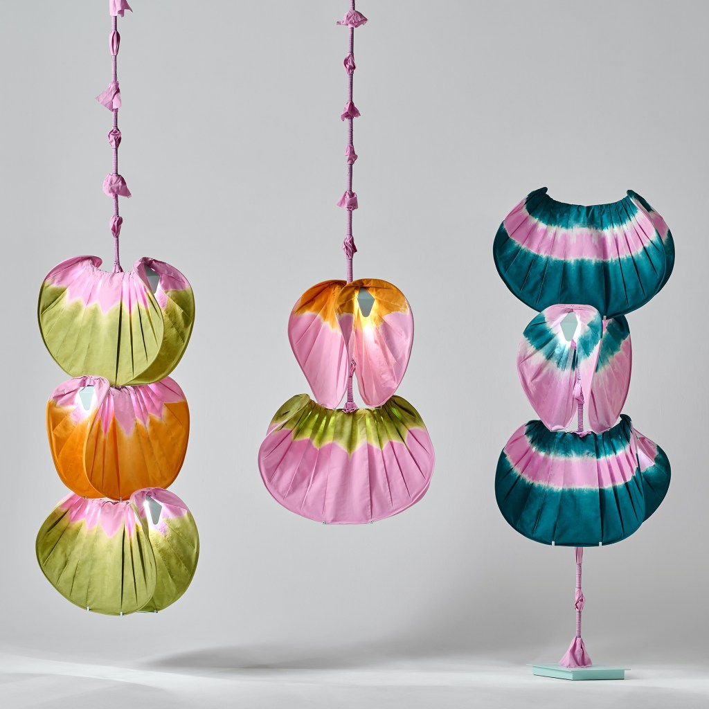 Three vibrant hanging fabric sculptures in shades of green, pink, orange, and blue, displayed against a light grey background.