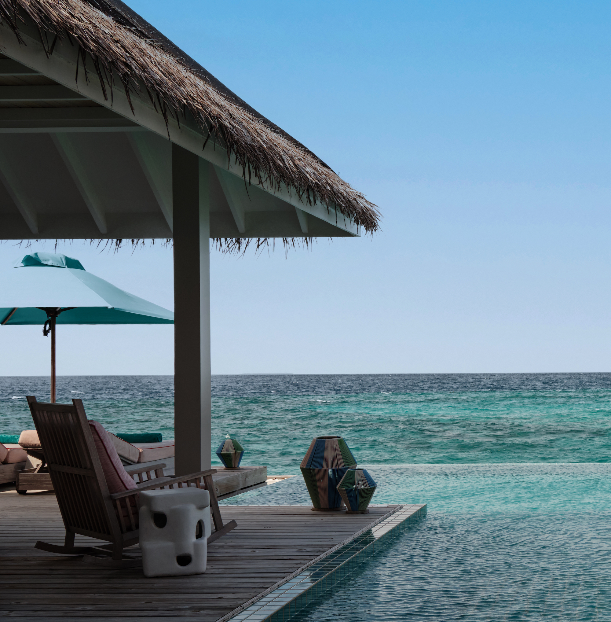 A view of a serene beachfront with turquoise water, featuring a wooden deck, a rocking chair, colourful lanterns, and a thatched roof shelter.